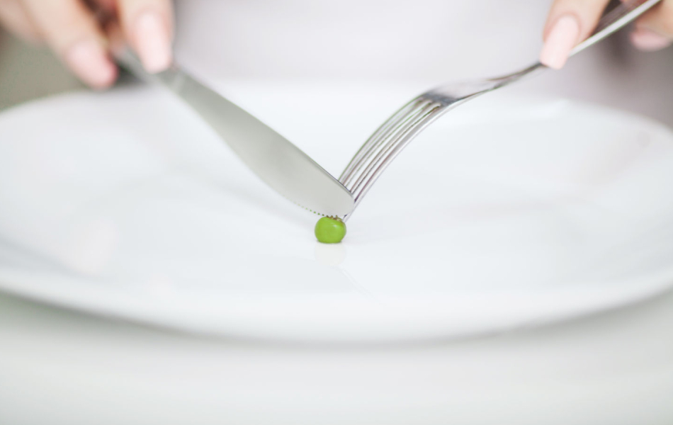 Fork and knife poised over a single green pea on a white plate.