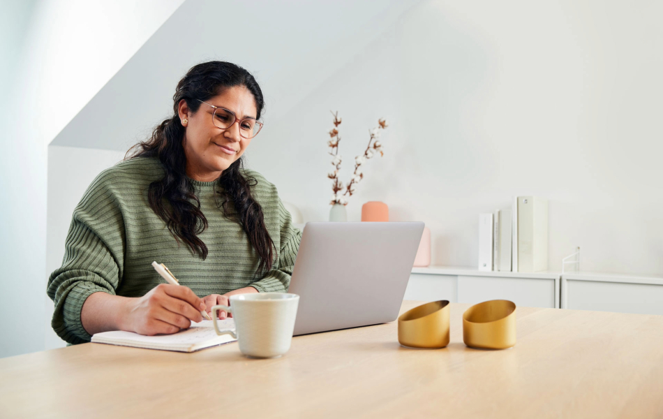 Person taking notes while using a laptop at a desk in a home office.