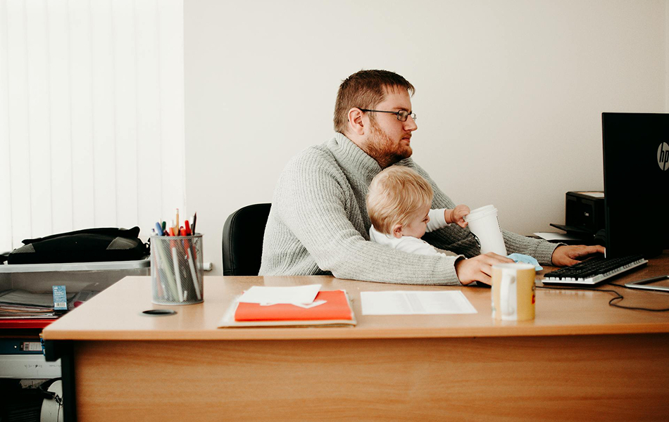 Person working at a computer in an office while holding a young child on their lap.
